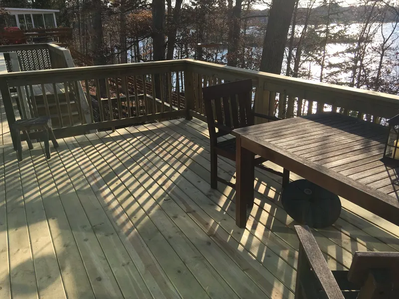 Pale wood deck with a railing, table and chairs, looking toward water.